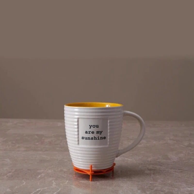 A white ribbed ceramic mug with a yellow interior and handle, featuring the printed phrase 'you are my sunshine' on the front, sitting on an orange coaster against a neutral background.