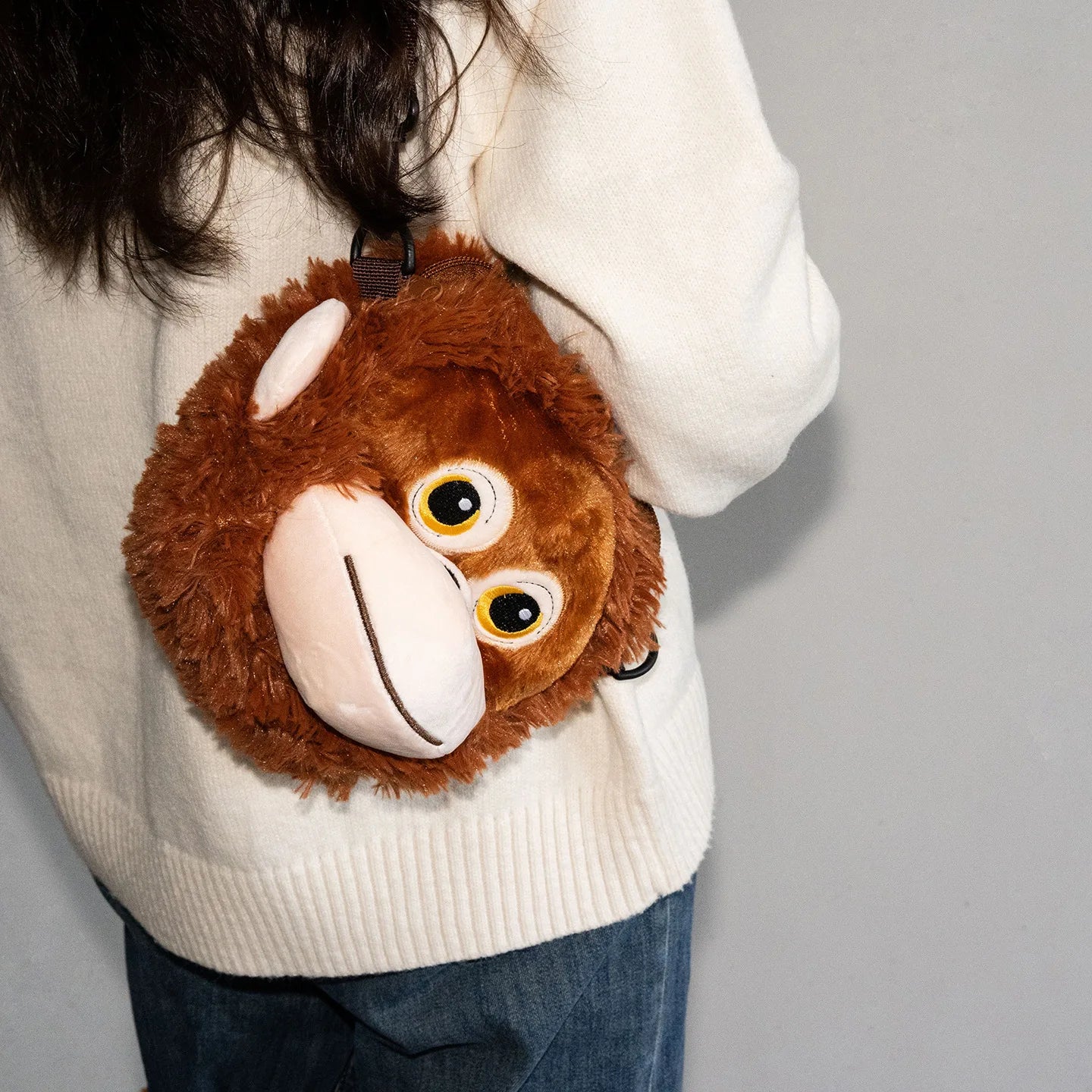Person wearing a white sweater with a brown plush monkey bag on a gray background