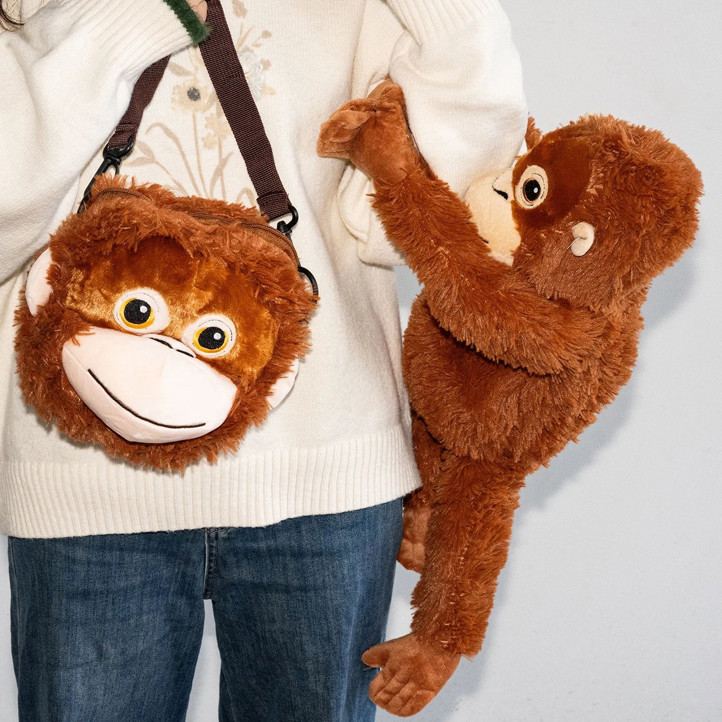 Person holding a plush monkey toy and a monkey-shaped bag against a light gray background