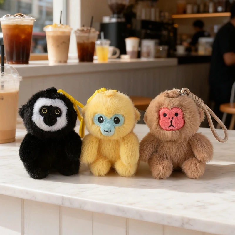Three plush monkey keychains on a marble counter with a coffee shop background.