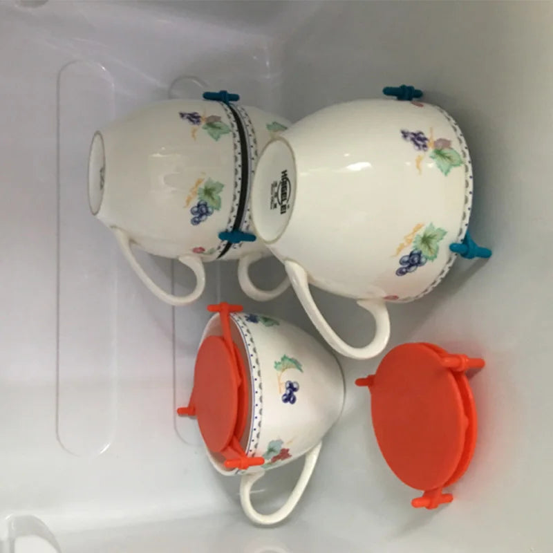 Three white ceramic teacups with floral grape prints and blue rubber grommets, shown upside down inside a clear plastic storage container, with two orange silicone lids placed beside them.