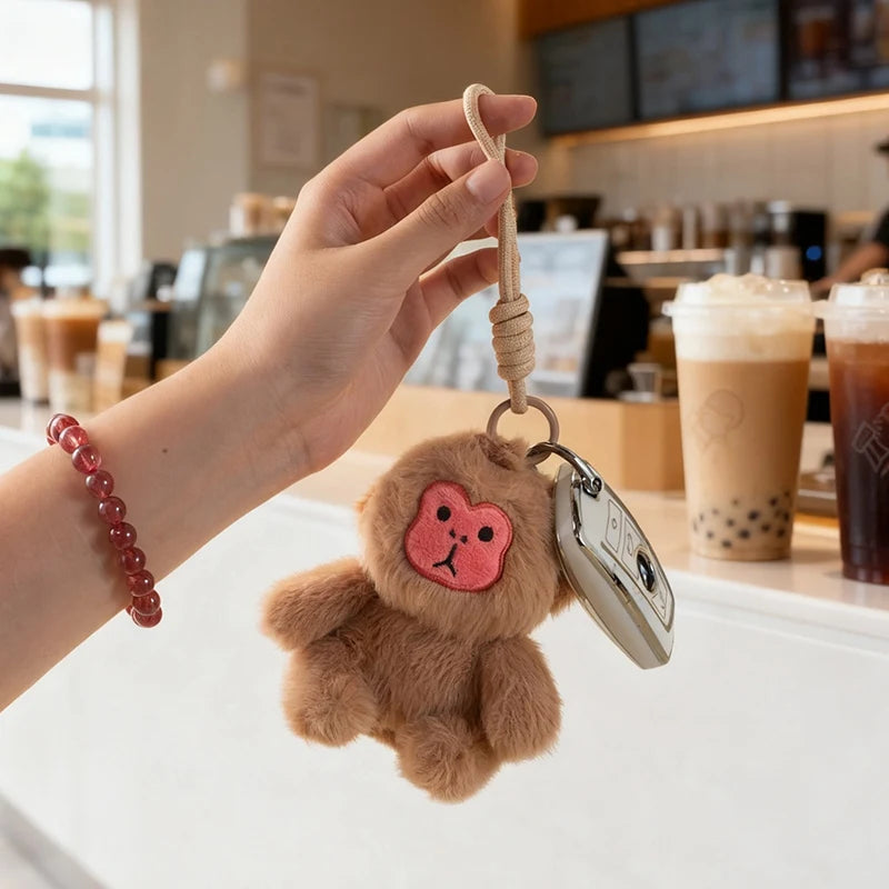 Brown plush monkey keychain with a pink face held by a hand in a coffee shop.